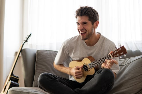 Happy young man is playing ukulele guitar and singing on the sofa at home