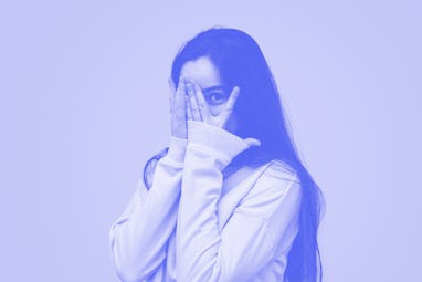 Timid teen girl with long hair covering face with hand and peeking through fingers against pink back...