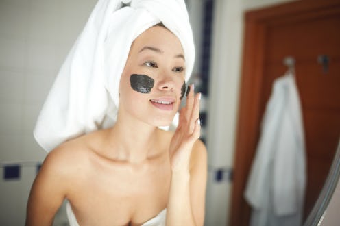 Portrait of beautiful Asian woman applying face mask during beauty treatment looking in mirror and s...