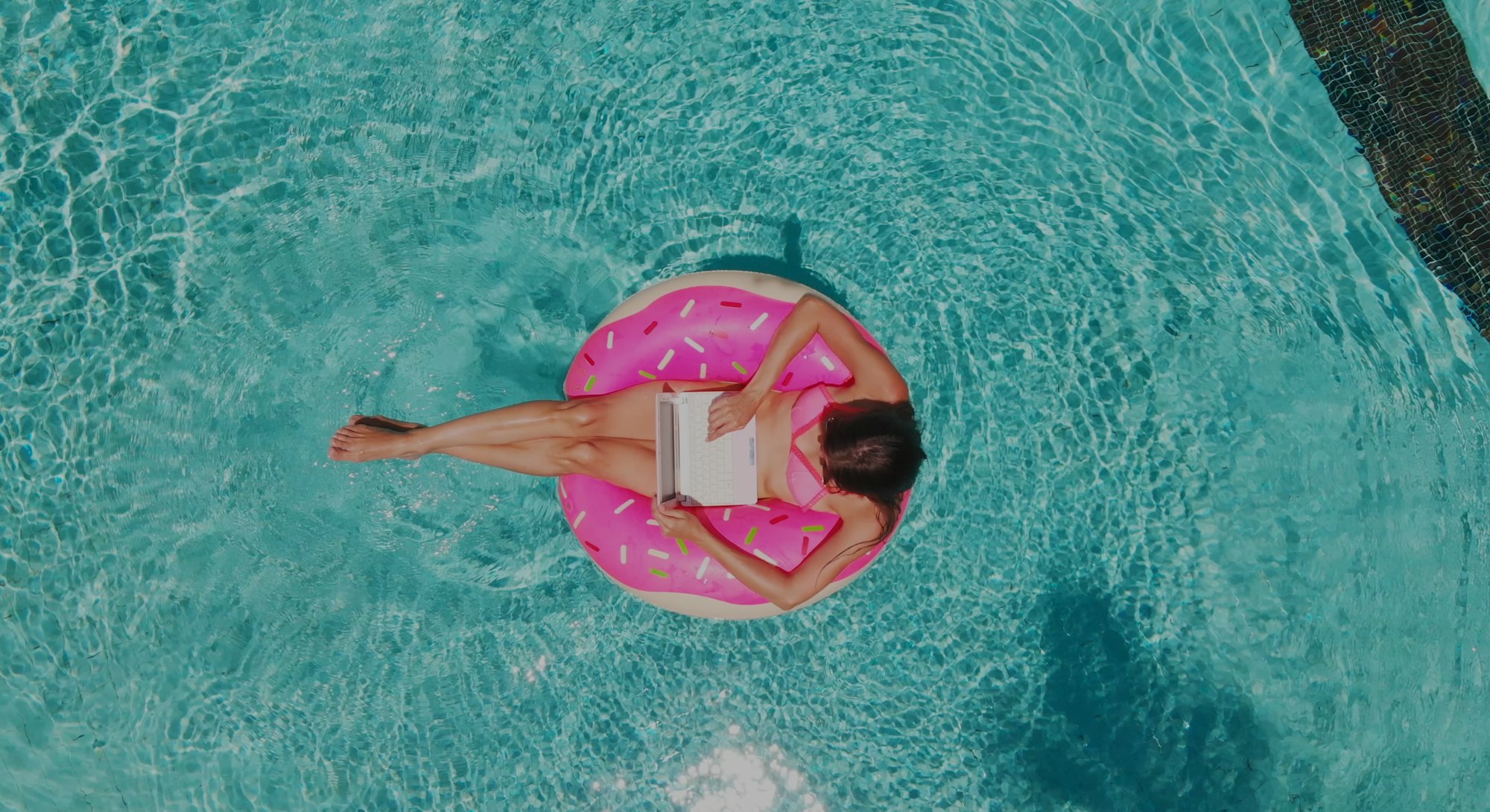 Aerial view of a young brunette woman swimming on an inflatable big donut with a laptop in a transpa...
