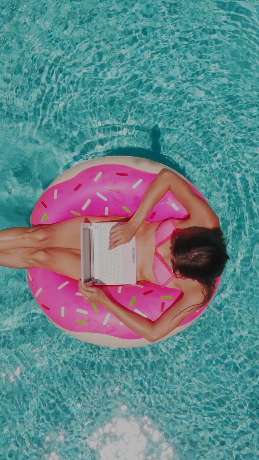 Aerial view of a young brunette woman swimming on an inflatable big donut with a laptop in a transpa...