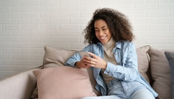Happy african millennial woman holding smart phone watching social media video content. Smiling youn...