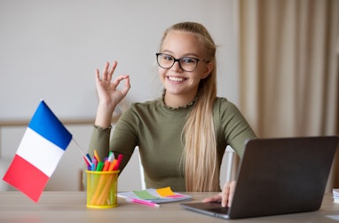 Cheerful girl teenager with flag of France showing okay gesture, using laptop, having educational on...