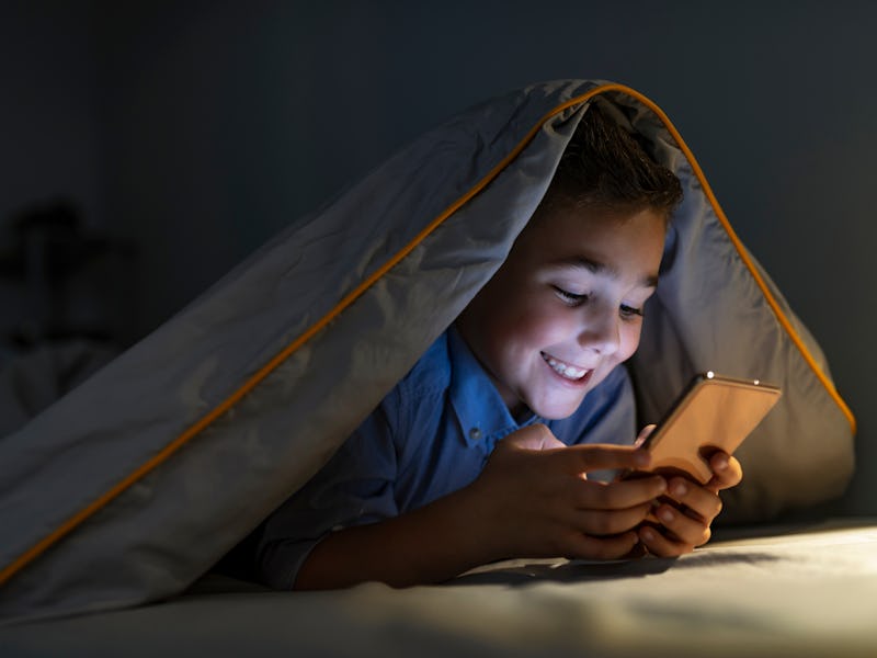 Kid boy playing smartphone lying on a bed at night. 7 years old child boy using smartphone at night ...