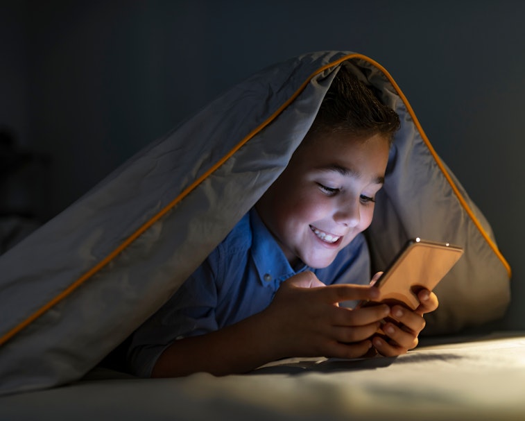 Kid boy playing smartphone lying on a bed at night. 7 years old child boy using smartphone at night ...