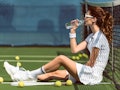 side view of beautiful tennis player in white tennis uniform and sunglasses drinking water while res...