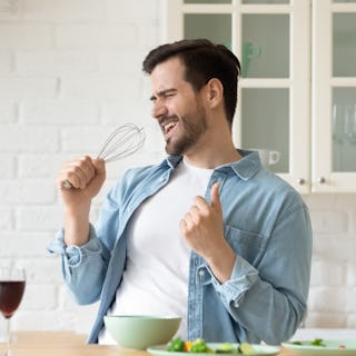 Funny guy singing song holding beater whisk microphone listening music in modern kitchen, happy joyf...