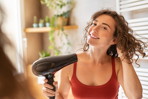 Beautiful girl using a hair dryer and smiling. Natural young woman drying curly hair with hair-dry m...