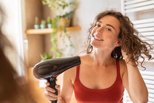 Beautiful girl using a hair dryer and smiling. Natural young woman drying curly hair with hair-dry m...