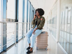 Portrait of happy young african american woman sitting on suitcase and talking with mobile phone at ...