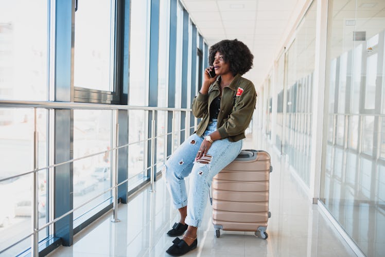 Portrait of happy young african american woman sitting on suitcase and talking with mobile phone at ...