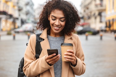 Young woman smiling while having coffee and reading her October 2021 monthly horoscope on her phone.