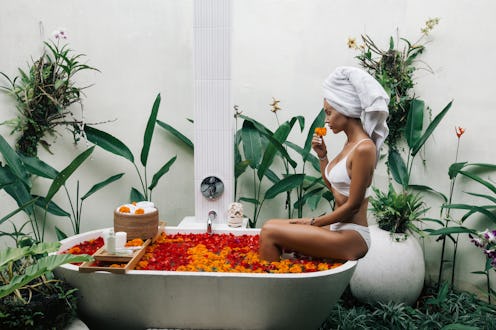Woman with wrapped towel on head is relaxing in outdoor bath with flowers in Bali spa hotel.