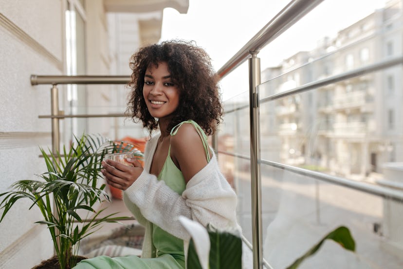 A woman smiles on an outdoor patio with plants around her, holding a beverage. Cusp signs in astrolo...