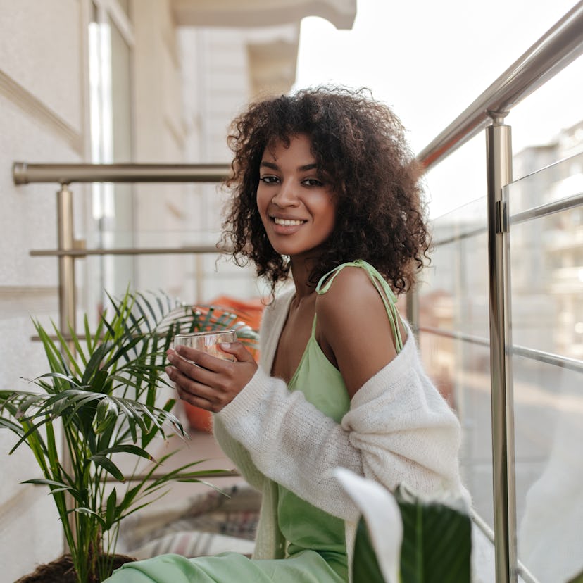 Happy brunette woman drinks coffee on balcony. Charming lady in light green dress smiles sincerely o...