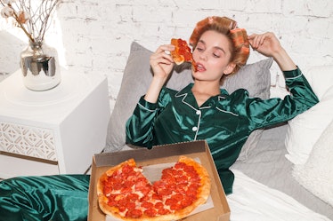 young fashionable woman in silk pajamas lays on bed and eat pizza pepperoni from pizza box, direct f...