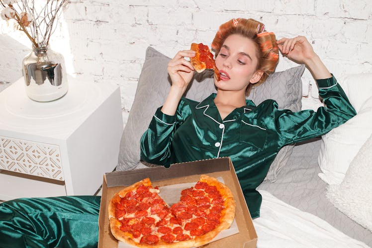 young fashionable woman in silk pajamas lays on bed and eat pizza pepperoni from pizza box, direct f...