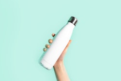 Close-up of female hand, holding white reusable steel stainless eco thermo water bottle with mockup,...