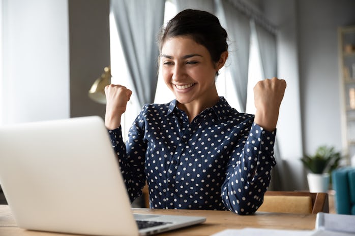 Overjoyed indian ethnicity girl sit at desk looks at laptop screen read incredible news clench fists...