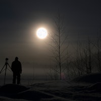 Photographer standing in snow photographing winter landscape. Full Moon and stars on night sky.