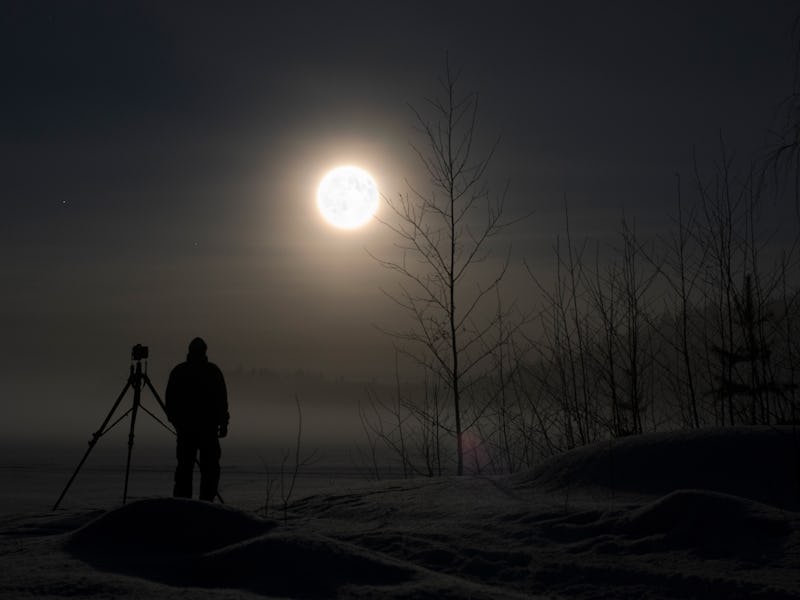 Photographer standing in snow photographing winter landscape. Full Moon and stars on night sky.