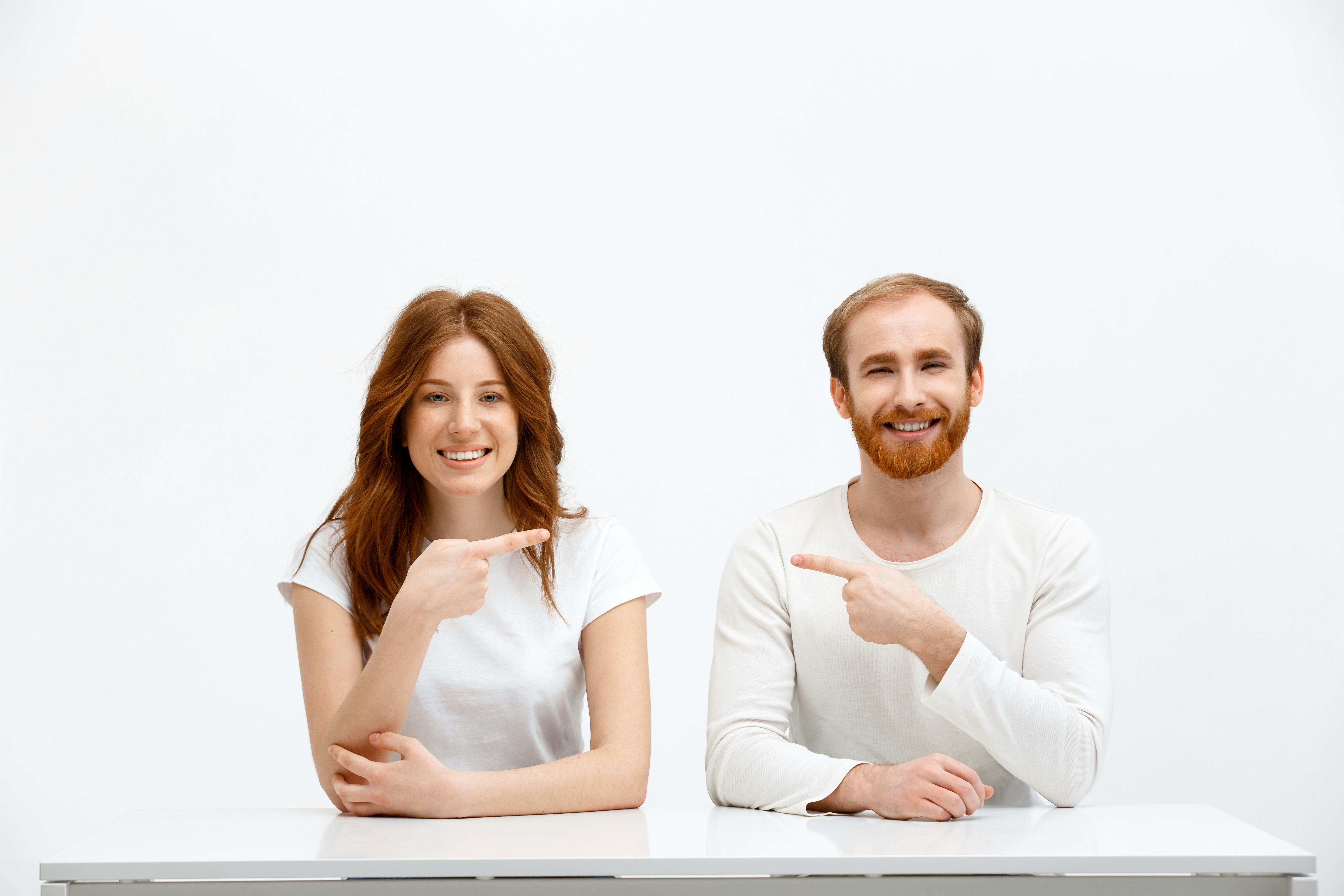 Funny redhead boy and girl sitting at white desk over white background and point finger to each othe...