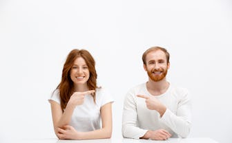Funny redhead boy and girl sitting at white desk over white background and point finger to each othe...