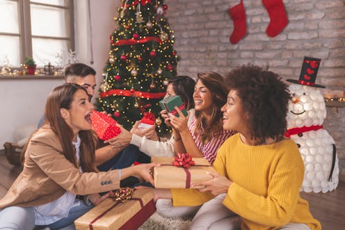 Group of friends sitting by nicely decorated Christmas tree, celebrating Christmas at home, exchangi...