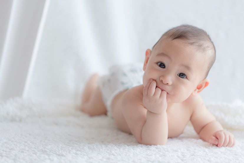 Asian baby boy lying on his tummy on a piece of white fluffy cloth chewing his hands and smiling at ...