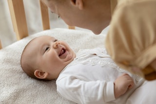 New mom talking to happy adorable few month baby resting in crib and smiling. Young mother speaking ...