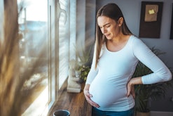 Woman experiencing stomach or pelvic pain during pregnancy stands near window