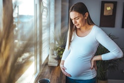 Woman experiencing stomach or pelvic pain during pregnancy stands near window