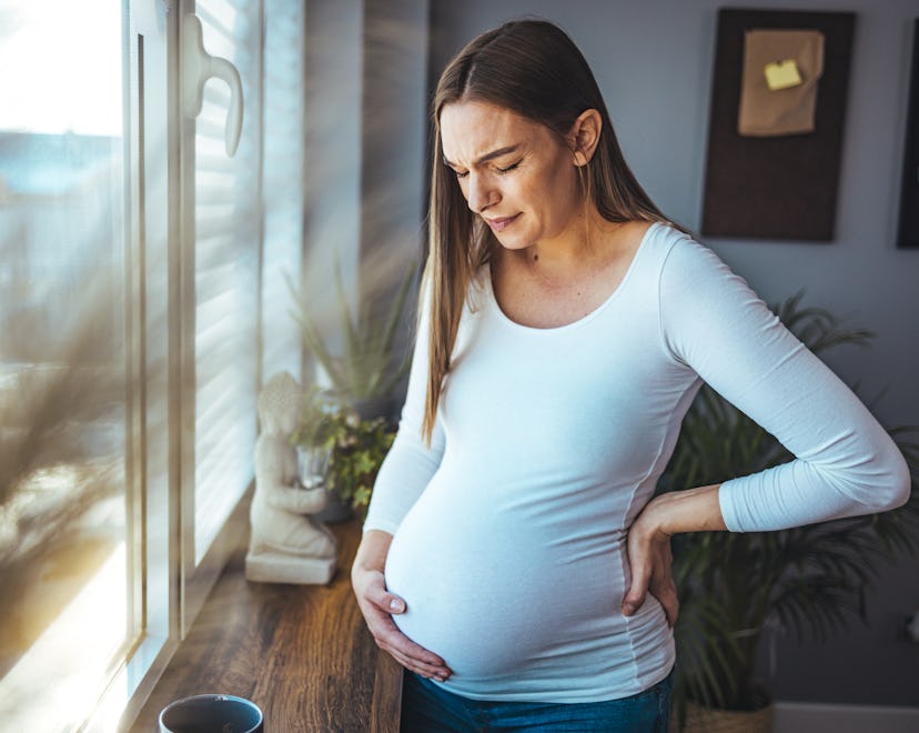 Woman experiencing stomach or pelvic pain during pregnancy stands near window