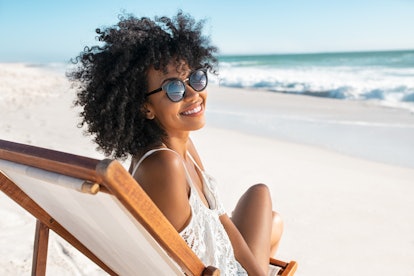 Portrait of happy young black woman relaxing on wooden deck chair at tropical beach while looking at...