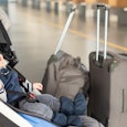 Cute funny caucasian baby boy sitting in stroller near luggage at airport terminal. Child sin carria...