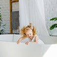 A little girl with light curly hair is laughing and sitting in the bathroom. Close-up portrait. Chil...