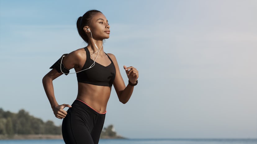 Side view shot of young woman in sportswear jogging on beach. African-american female jogger runner ...