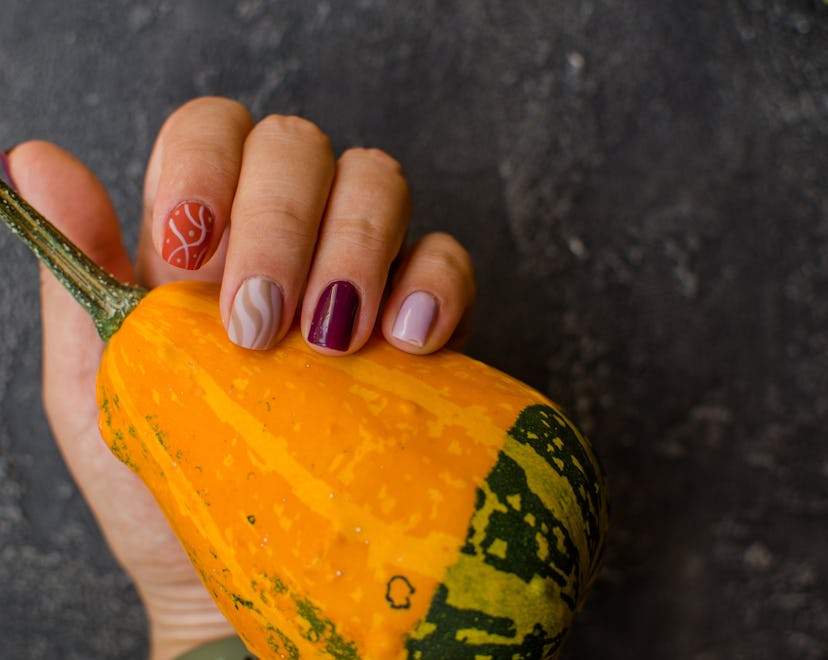 A Thanksgiving nail design with maroon, gold, orange, and swirls on a manicured hand holding a small...