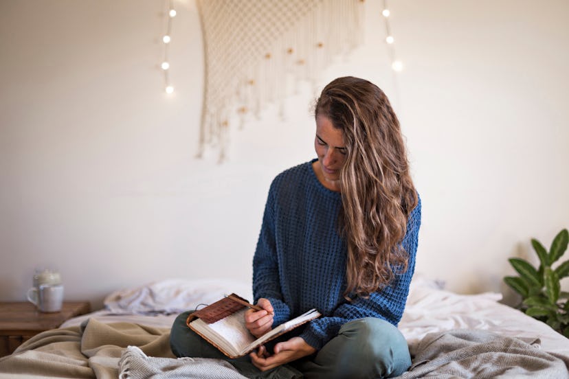 Woman in blue sweater sat on her bed writing in her leatherbound journal