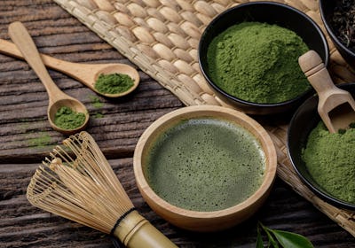 Japanese organic matcha green tea powder in bowl with wire whisk and green tea leaf on wooden backgr...