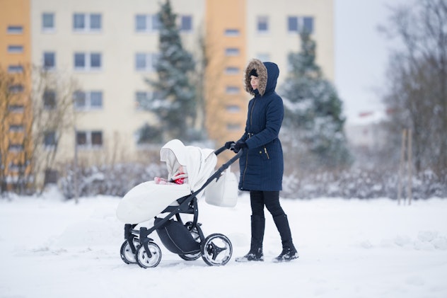 Young adult mother pushing white baby stroller and walking on snow covered sidewalk at town after bl...