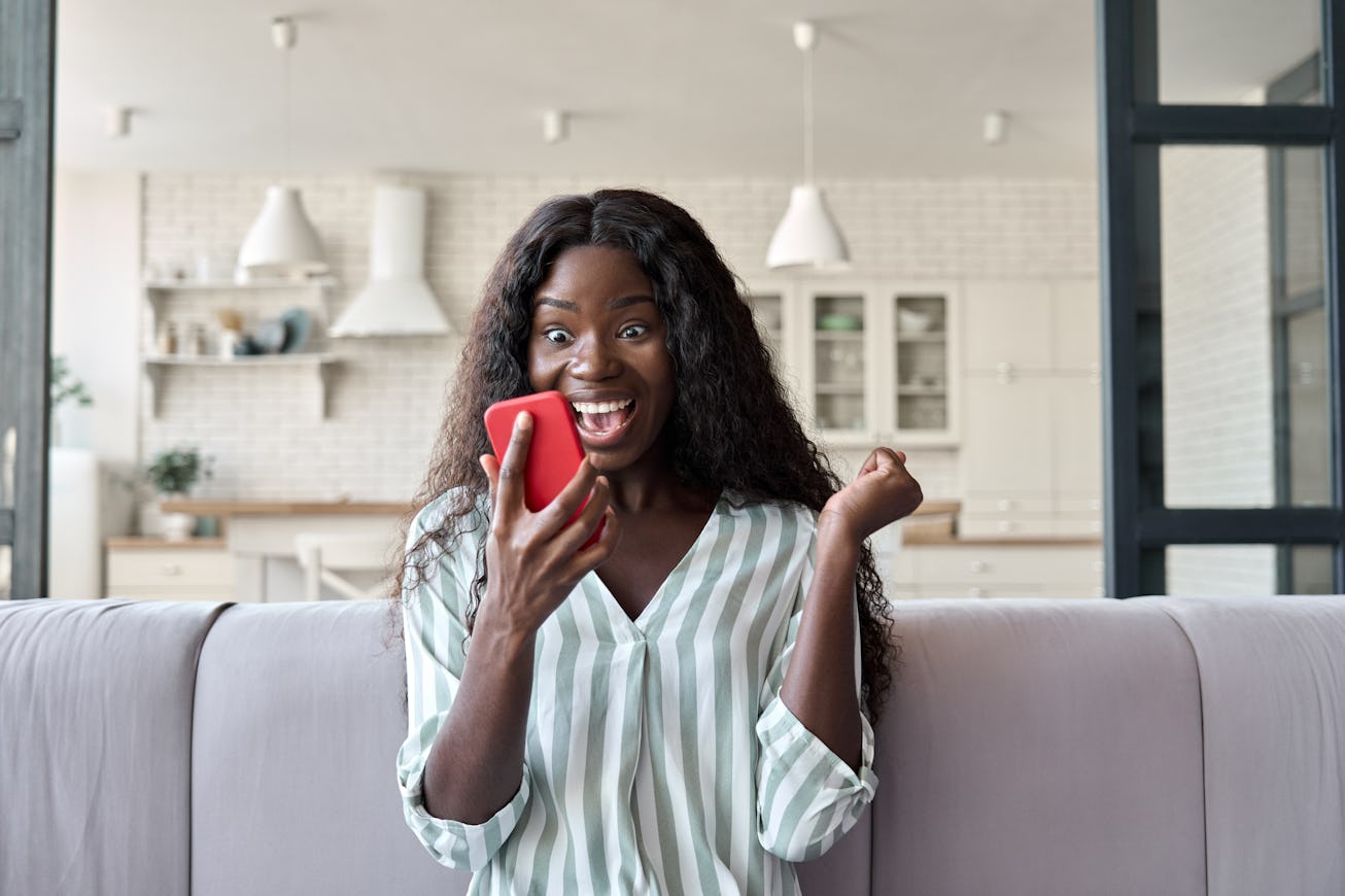 Young African girl at home reading message on mobile phone on unexpected news, happy client winning ...