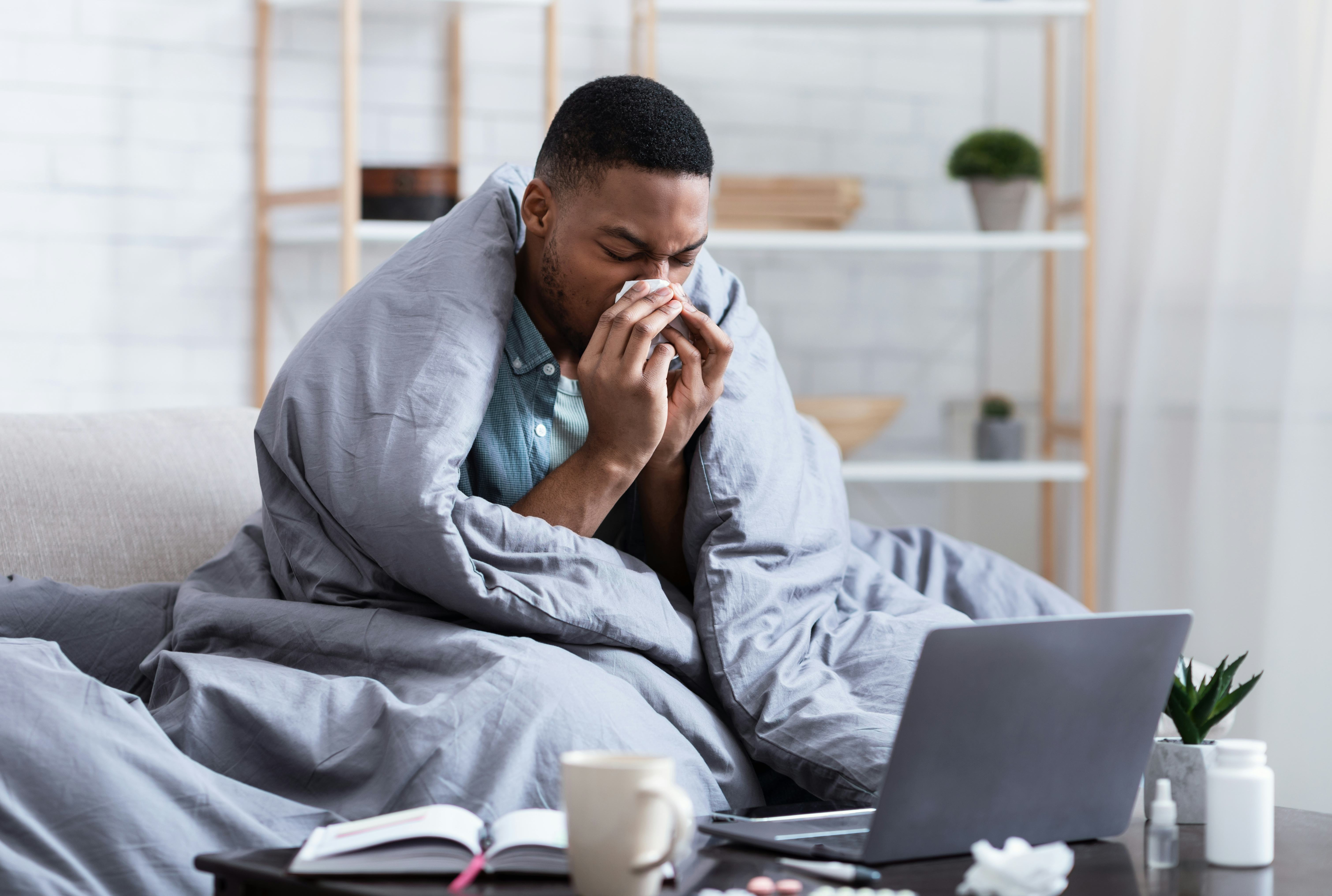 Ill African Man Having Rhinitis Sneezing And Blowing Runny Nose In Paper Tissue Sitting On Sofa At H...