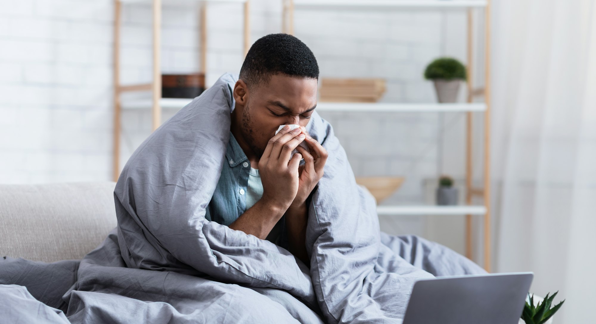 Ill African Man Having Rhinitis Sneezing And Blowing Runny Nose In Paper Tissue Sitting On Sofa At H...