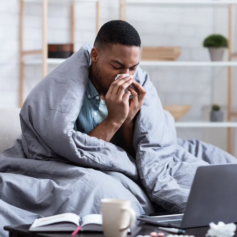 Ill African Man Having Rhinitis Sneezing And Blowing Runny Nose In Paper Tissue Sitting On Sofa At H...