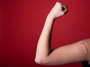 A girl makes a fist and flexes her bicep in front of a red background while wearing a bandage with h...