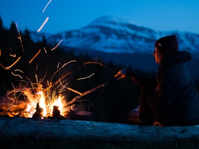 Girl relaxes near a bright campfire with sparks. Forest and snow-capped mountain in the background. ...