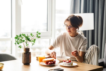 Photo of young serious woman eating fruits and pancakes while having breakfast in cozy room at home