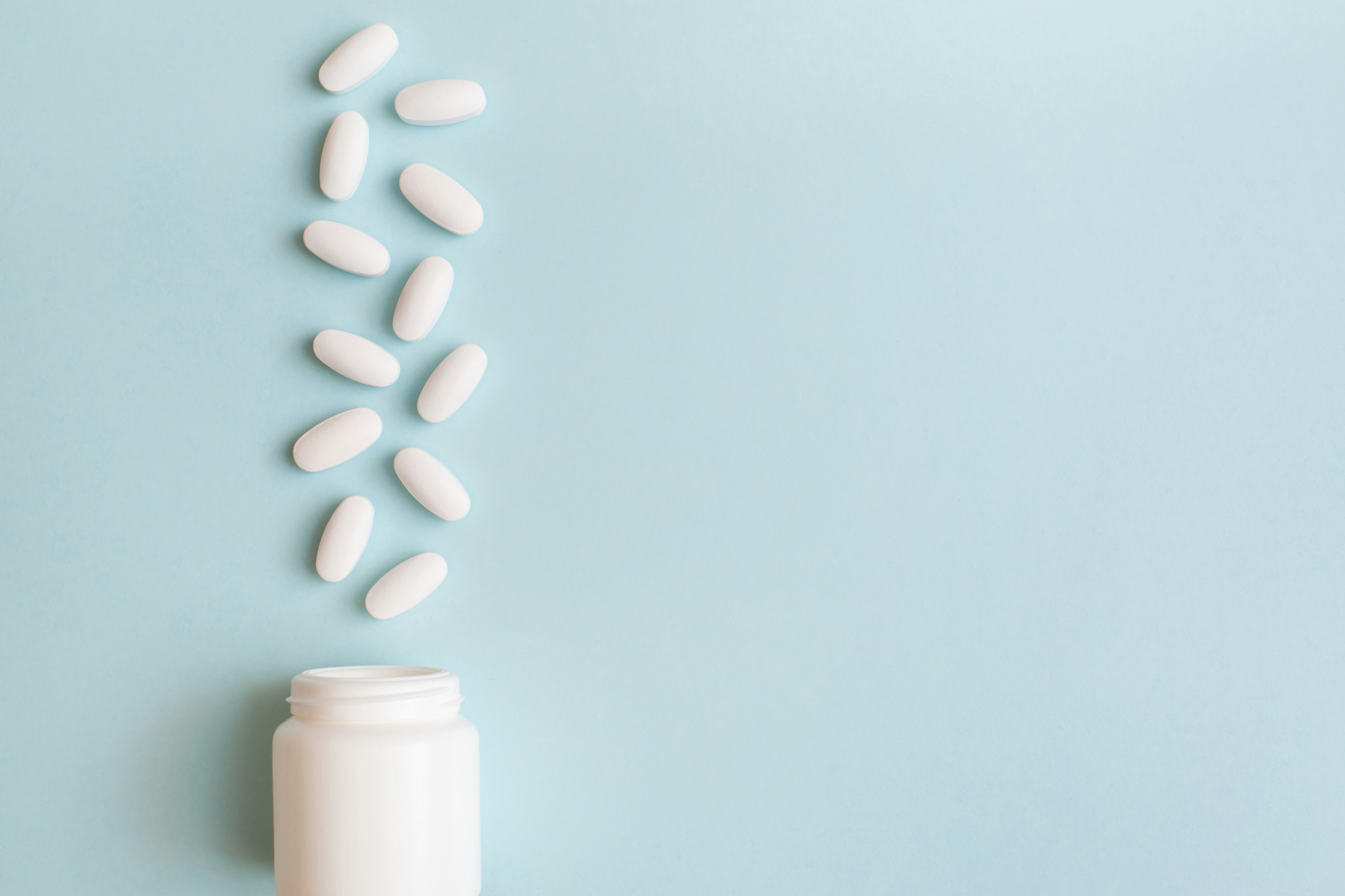 White pills and capsules spilled out of white bottle on light blue background. Minimal medical conce...