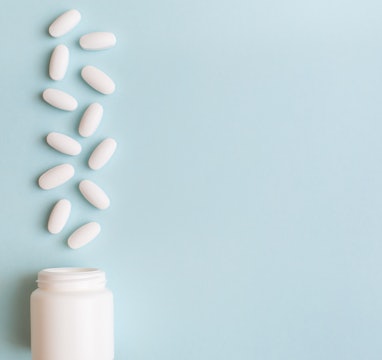 White pills and capsules spilled out of white bottle on light blue background. Minimal medical conce...
